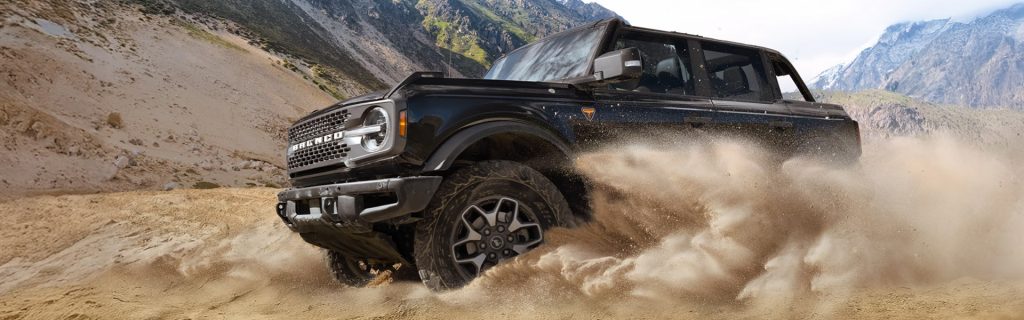 A black Ford Bronco off-road SUV kick up a large cloud of sand while driving through a rugged, mountainous desert terrain.