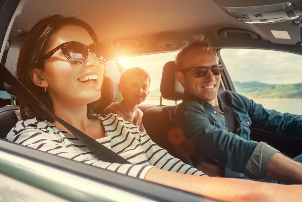 A happy family with a young boy in the back seat smiling and wearing sunglasses while on a sunny road trip inside their car.
